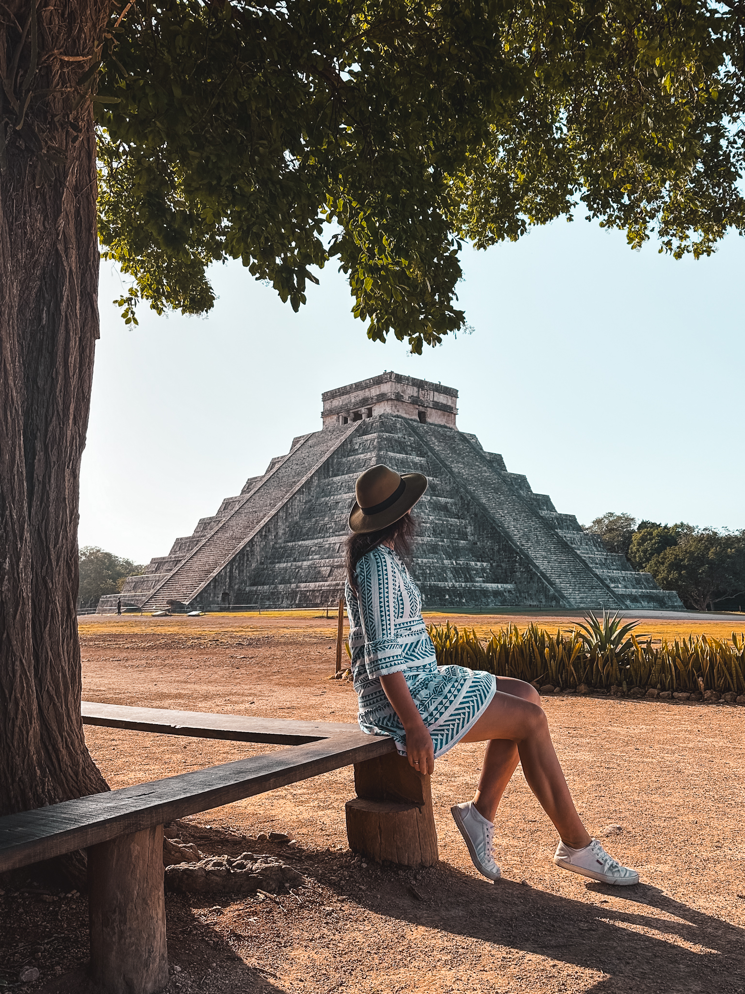 Valladolid, Mexico-best Instagram spots in Valladolid - Chichen Itza archaeological site - wooden bench under a lush tree with the Temple of Kukulkan Mayan pyramid in the background