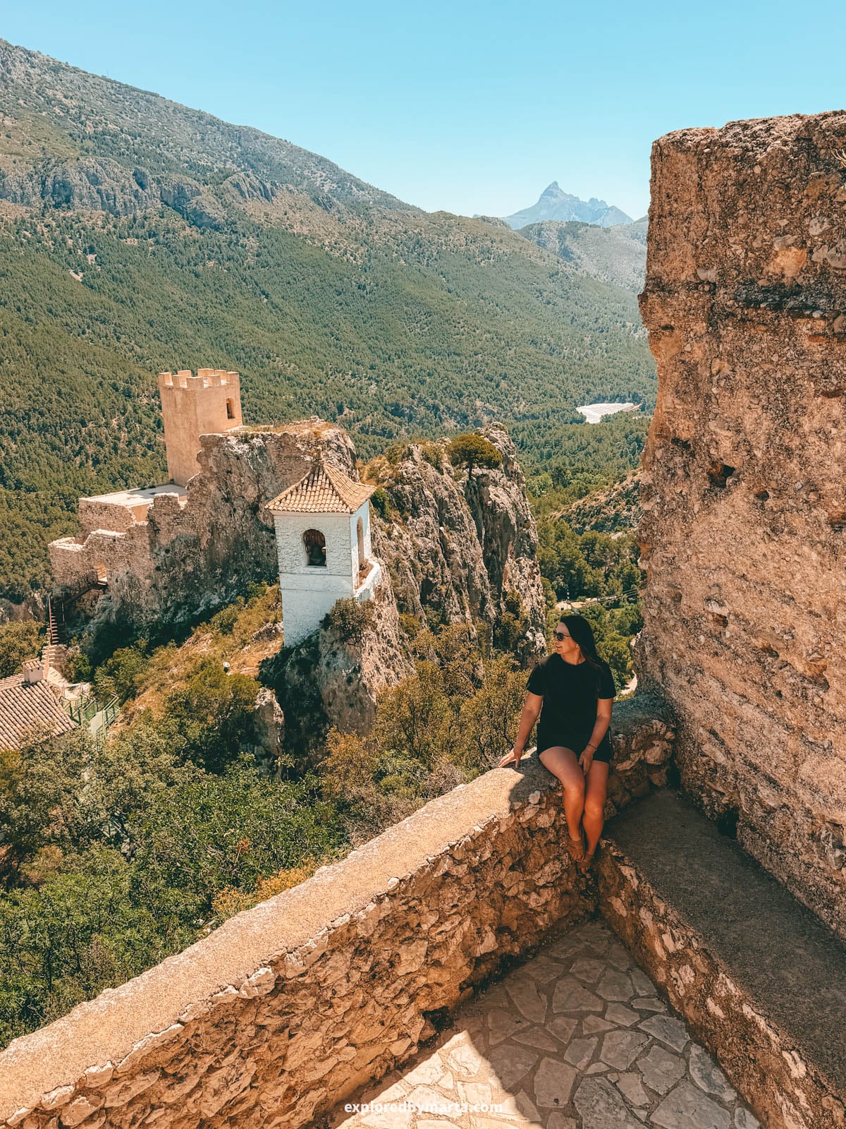 Sitting on the wall at San José castle with the Guadalest bell tower behind in Guadalest, Spain